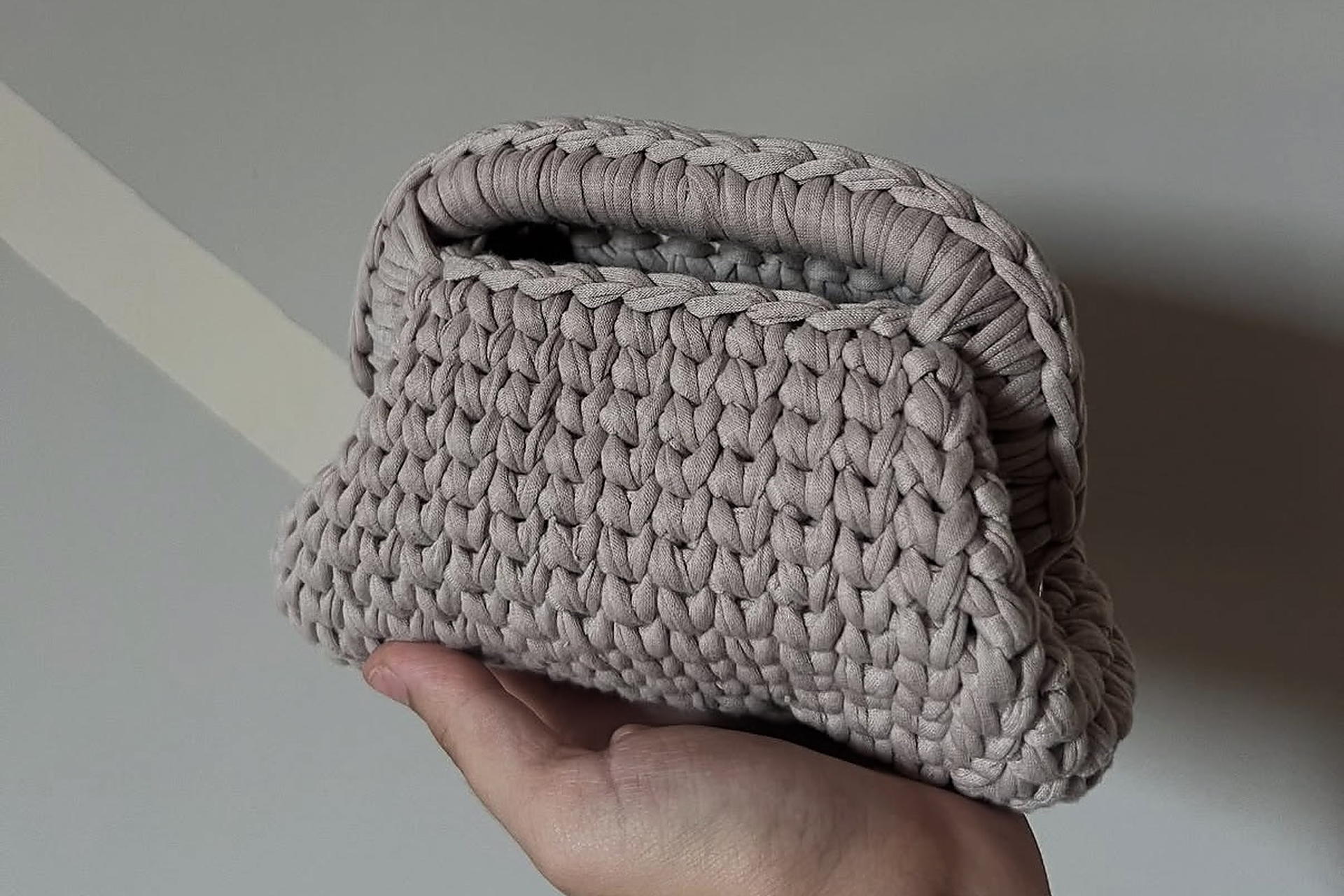 La pouch Nuvola in filato tortora, lavorata a mano, tenuta in mano davanti a una parete chiara